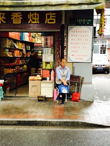 A street-side shop with an open front showcases a variety of colorful packaged goods inside. A man is seated at the entrance, holding a blue bag, with additional items like a red bag and cardboard boxes nearby. The storefront has signage with Chinese characters, and the surrounding environment appears to be urban with a road in the foreground.