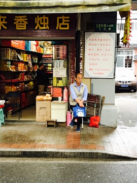 A street-side shop with an open front showcases a variety of colorful packaged goods inside. A man is seated at the entrance, holding a blue bag, with additional items like a red bag and cardboard boxes nearby. The storefront has signage with Chinese characters, and the surrounding environment appears to be urban with a road in the foreground.