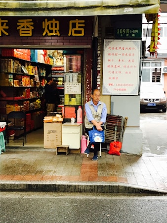 A street-side shop with an open front showcases a variety of colorful packaged goods inside. A man is seated at the entrance, holding a blue bag, with additional items like a red bag and cardboard boxes nearby. The storefront has signage with Chinese characters, and the surrounding environment appears to be urban with a road in the foreground.