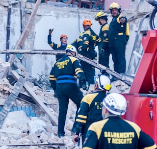 Rescue workers wearing helmets and uniforms are navigating a scene of destruction, likely from a collapsed building. They are surrounded by rubble and debris as they communicate and coordinate the rescue efforts.