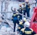 Rescue workers wearing helmets and uniforms are navigating a scene of destruction, likely from a collapsed building. They are surrounded by rubble and debris as they communicate and coordinate the rescue efforts.