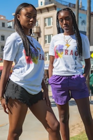 Two young individuals are posing confidently outdoors, wearing matching white t-shirts with colorful 'COURAGE' text and symbols. They are standing on a sunny day, with a background featuring buildings and palm trees. One person is wearing black shorts while the other is wearing purple shorts. They both have long hair styled differently.