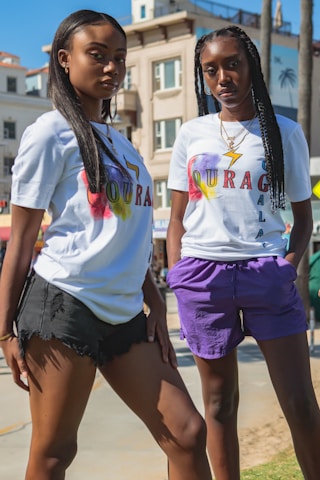 Two young individuals are posing confidently outdoors, wearing matching white t-shirts with colorful 'COURAGE' text and symbols. They are standing on a sunny day, with a background featuring buildings and palm trees. One person is wearing black shorts while the other is wearing purple shorts. They both have long hair styled differently.