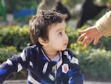 A young child with curly hair looks to the side, appearing curious or intrigued. The child is wearing a dark blue jacket with white and blue patterns, and there is a badge on the chest with a number. An adult hand reaches towards the child from the right. In the background, there is greenery suggesting a park or garden setting.