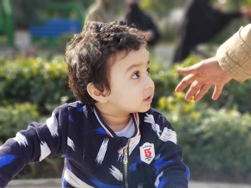 A young child with curly hair looks to the side, appearing curious or intrigued. The child is wearing a dark blue jacket with white and blue patterns, and there is a badge on the chest with a number. An adult hand reaches towards the child from the right. In the background, there is greenery suggesting a park or garden setting.
