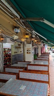 A row of outdoor dining tables with intricate mosaic patterns is lined up beneath a green awning. Above the tables, decorative lanterns in vibrant colors are suspended, adding an ornate touch. The setting gives a sense of a cafe or restaurant with a Middle Eastern or Mediterranean style. A display of colorful ceramics is visible on the wall to the left.