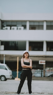 A person stands confidently with arms crossed in front of a building. The background shows a modern structure with large windows and air conditioning units. A white van is parked to the side on a paved surface.