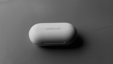 A white, oval-shaped OnePlus branded earbuds case placed on a smooth, dark surface. The lighting creates a slight shadow beneath the case, suggesting a soft, diffused light source.