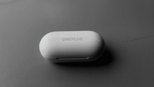 A white, oval-shaped OnePlus branded earbuds case placed on a smooth, dark surface. The lighting creates a slight shadow beneath the case, suggesting a soft, diffused light source.