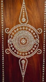 Intricate geometric patterns and designs are created using white inlays on a dark brown wooden surface. The central design features circular and triangular shapes with a floral motif in the center, bordered by repetitive triangular patterns.