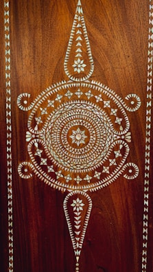 Intricate geometric patterns and designs are created using white inlays on a dark brown wooden surface. The central design features circular and triangular shapes with a floral motif in the center, bordered by repetitive triangular patterns.