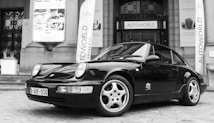 A sleek black classic sports car is parked in front of an entrance to an exhibition hall. The building features large columns and banners with 'Autoworld' text. The cobblestone pavement adds to the historical ambiance of the scene. Light reflects off the car's polished surface, enhancing its contours and design.