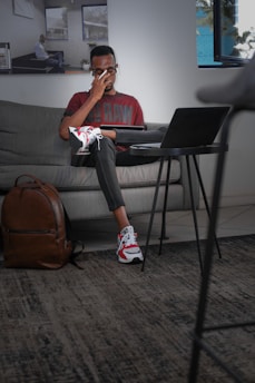 A person sits on a gray sofa, wearing glasses and using a laptop on a small table. The individual has their hand on their forehead, possibly indicating stress or focus. A brown backpack rests on the floor nearby. The room has a modern interior with framed pictures on the wall and a window letting in natural light.