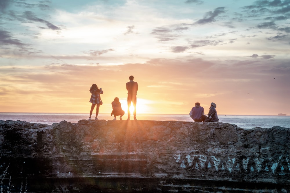 silhouette of 3 people standing on rock formation near body of water during sunset