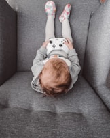 A child sitting on a gray couch holding a white video game controller. The child is wearing gray and white clothing with colorful patterned slippers.