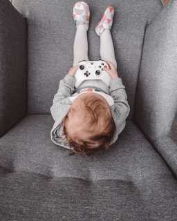 A child sitting on a gray couch holding a white video game controller. The child is wearing gray and white clothing with colorful patterned slippers.