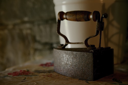 An old, rustic iron with a wooden handle rests on a patterned surface in a dimly lit setting. The iron's metal shows signs of rust and age, while the background is a soft focus, adding to the vintage feel of the image.