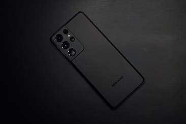 A black smartphone placed on a dark, textured surface. The device features multiple camera lenses and sensors on the top left corner of the back panel, with the brand name 'Samsung' visible near the bottom.