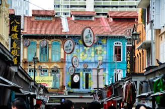 The vibrant urban street features colorful shophouses with intricate wooden windows and red-tiled roofs. Hanging circular signs with Chinese characters are interspersed with traditional lanterns. The scene includes a busy street market below, with stalls lined up and a variety of goods displayed. A few people are visible walking around.