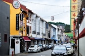 A vibrant urban street lined with colorful shophouses featuring various signs and advertisements, including those for Thai food and kitchenware. Cars are parked along the side, and a few pedestrians are visible. Decorative lights and signage hang above the street, adding to the lively atmosphere.