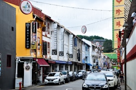 A vibrant urban street lined with colorful shophouses featuring various signs and advertisements, including those for Thai food and kitchenware. Cars are parked along the side, and a few pedestrians are visible. Decorative lights and signage hang above the street, adding to the lively atmosphere.