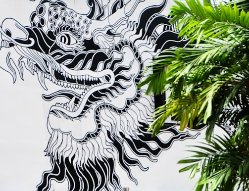 A monochrome mural depicts a stylized dragon with intricate detailing, featuring bold black outlines and patterns. The dragon's fierce expression is accentuated by sharp teeth and swirling shapes. The mural is partially obscured by the lush green fronds of a palm tree on the right.