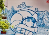 A mural featuring a large blue line-art illustration of a person wearing a helmet, with a serene expression, surrounded by stylized nature elements like clouds, trees, and a landscape with water and rocks. The art is painted on a large wall, incorporating real windows and surrounding greenery.