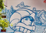 A mural featuring a large blue line-art illustration of a person wearing a helmet, with a serene expression, surrounded by stylized nature elements like clouds, trees, and a landscape with water and rocks. The art is painted on a large wall, incorporating real windows and surrounding greenery.