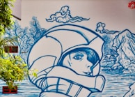 A mural featuring a large blue line-art illustration of a person wearing a helmet, with a serene expression, surrounded by stylized nature elements like clouds, trees, and a landscape with water and rocks. The art is painted on a large wall, incorporating real windows and surrounding greenery.