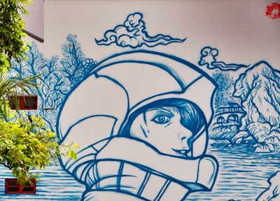 A mural featuring a large blue line-art illustration of a person wearing a helmet, with a serene expression, surrounded by stylized nature elements like clouds, trees, and a landscape with water and rocks. The art is painted on a large wall, incorporating real windows and surrounding greenery.