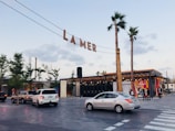 An outdoor scene features a shopping and entertainment area with the sign 'LA MER' suspended overhead. The environment includes palm trees, modern architecture, and several parked cars. People walk near a building with large colorful wall art featuring abstract designs.