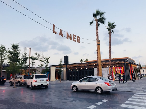 An outdoor scene features a shopping and entertainment area with the sign 'LA MER' suspended overhead. The environment includes palm trees, modern architecture, and several parked cars. People walk near a building with large colorful wall art featuring abstract designs.