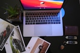A laptop with a vivid red and orange screen sits on a dark surface. Surrounding the laptop are glossy car brochures, a succulent in a square pot, a leather wallet, and a set of car keys with a distinctive logo.