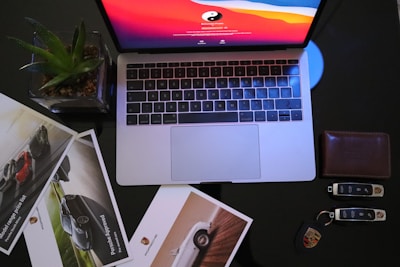 A laptop with a vivid red and orange screen sits on a dark surface. Surrounding the laptop are glossy car brochures, a succulent in a square pot, a leather wallet, and a set of car keys with a distinctive logo.