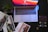 A laptop with a vivid red and orange screen sits on a dark surface. Surrounding the laptop are glossy car brochures, a succulent in a square pot, a leather wallet, and a set of car keys with a distinctive logo.