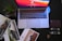 A laptop with a vivid red and orange screen sits on a dark surface. Surrounding the laptop are glossy car brochures, a succulent in a square pot, a leather wallet, and a set of car keys with a distinctive logo.
