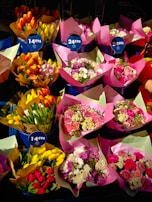 A variety of flower bouquets are arranged in buckets, each wrapped in colorful paper. The bouquets contain a mix of tulips, roses, and daisies, with some brightly colored papers and tags indicating prices.