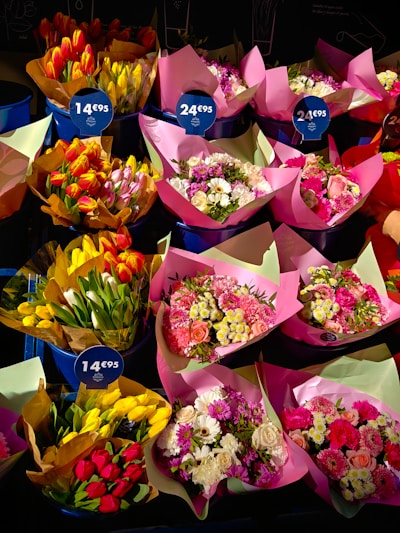 A variety of flower bouquets are arranged in buckets, each wrapped in colorful paper. The bouquets contain a mix of tulips, roses, and daisies, with some brightly colored papers and tags indicating prices.
