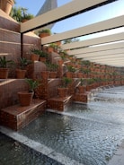Terraced brick planters filled with small green plants are arranged along a water feature with streams of water cascading down. The plant pots are evenly spaced, creating a pattern under a structure with beams providing partial shade. The setting is serene, with a mix of natural and architectural elements.