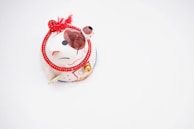 A small, decorative figurine of a cat with a white body, red and black accents, and a red rope tied around its neck. The cat features a cheerful expression and a paw raised in a beckoning gesture. A small bell is attached to the red rope, adding a touch of charm.