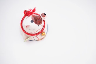 A small, decorative figurine of a cat with a white body, red and black accents, and a red rope tied around its neck. The cat features a cheerful expression and a paw raised in a beckoning gesture. A small bell is attached to the red rope, adding a touch of charm.