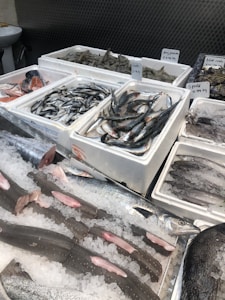 Various types of fresh seafood are displayed on ice in a market setting. Fish, shrimp, squid, and blue crabs are neatly arranged in white styrofoam boxes, with each type labeled with price tags. The scene reflects a typical seafood market with a variety of options available for purchase.