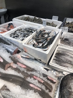 Various types of fresh seafood are displayed on ice in a market setting. Fish, shrimp, squid, and blue crabs are neatly arranged in white styrofoam boxes, with each type labeled with price tags. The scene reflects a typical seafood market with a variety of options available for purchase.