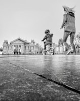 A black and white image features a child on a bicycle, wearing a helmet, in motion across a pavement. An adult, possibly a parent, is walking alongside the child. In the background, there is a large, classical-style building with prominent columns and ornate features.