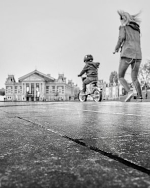 A black and white image features a child on a bicycle, wearing a helmet, in motion across a pavement. An adult, possibly a parent, is walking alongside the child. In the background, there is a large, classical-style building with prominent columns and ornate features.