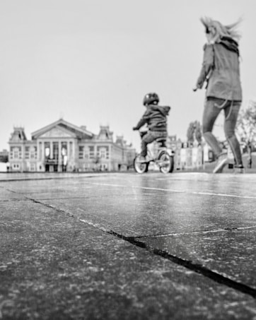A black and white image features a child on a bicycle, wearing a helmet, in motion across a pavement. An adult, possibly a parent, is walking alongside the child. In the background, there is a large, classical-style building with prominent columns and ornate features.