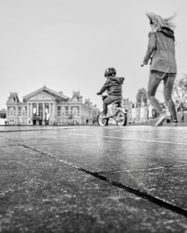 A black and white image features a child on a bicycle, wearing a helmet, in motion across a pavement. An adult, possibly a parent, is walking alongside the child. In the background, there is a large, classical-style building with prominent columns and ornate features.
