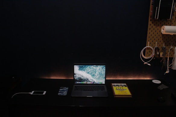 A minimalist workspace setup on a dark desk featuring a laptop with a screensaver of ocean waves. The setup includes a smartphone, a book with a bright yellow cover, and cable organizers on a pegboard attached to the wall. The ambient lighting creates a calm atmosphere.