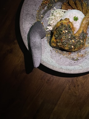 A meal is served on a large stone pestle and mortar. The dish includes fried chicken with a green seasoning or sauce, a serving of steamed white rice topped with a small garnish, shredded cabbage, and slices of tempeh. The meal is presented in a rustic style, with the stoneware providing a natural, earthy background. The lighting is warm and focused, creating shadows and highlights on the food.