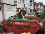 A residential street scene with a row of semi-detached houses. In the foreground, a large, red skip bin filled with construction debris, including wood, dirt, a green tarp, and patterned fabric, is situated on a driveway. Two cars are parked further back in front of the houses, which have a mix of decorative facades and bay windows.